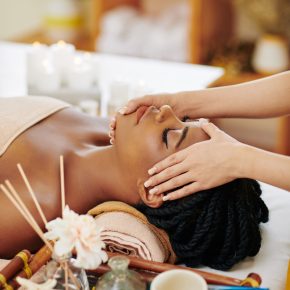 Beautiful young Black woman enjoying face and head massage in spa salon when lying on bed next to tray with aroma oils